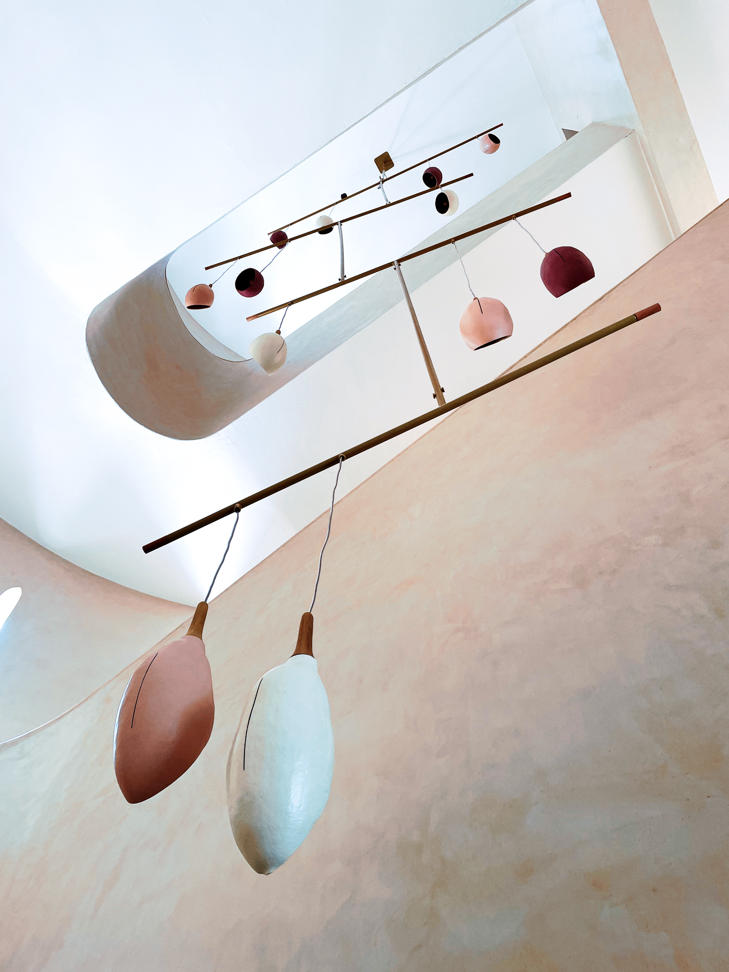 Seedling Designs bespoke installation. A modern mobile and pendant light fixture suspended from a ceiling in a minimalist interior. The mobile features a long horizontal brass bar with small geometric weights in pink, white, and dark red, with two larger organic-shaped pendant shades below in dusty pink and off-white.